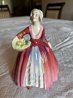 Front diagonal view of the Royal Doulton porcelain figurine depicting a woman holding a basket of flowers, wearing a red-pink dress with white apron and bonnet.
