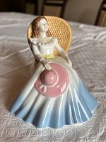 Front and angled view of the Royal Doulton 'Annabel' porcelain figurine seated on a wicker chair, dressed in a white ruffled top and skirt with blue shading holding a pink hat.