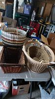 Photo showing four baskets on a plastic table in a storage or garage environment. Three woven baskets with handles and one beige plastic laundry basket.