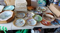 Overview of 20 assorted collector's plates displayed on a table, varying in themes including flowers, animals, children, boats, and scenic views.