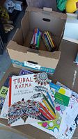 Cardboard box containing multiple sets of assorted markers and pencil crayons. Partially completed colouring books shown beside the box including Tribal Karma book with some pages coloured.
