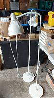 Two white floor lamps with adjustable arms and conical shades, standing side by side with cords and switches visible.
