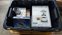 Photo of lot items inside a black storage tote showing boxed and loose items: Black & Decker food processor manual and base, Star frit electronic kitchen scale, Wilton cookie press in box.