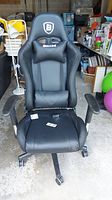 Front view of the black faux leather gaming chair showing overall shape, seat, armrests, and base with wheels. Remote controls laid on seat.