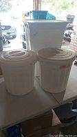 Two white plastic garbage pails with lids and one tall white plastic laundry hamper with attached lid on a table.