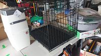 Photo shows the metal collapsible wire pet crate next to a white and black LitterLocker cat litter disposal bin and various pet supplies on table.