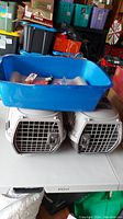 Two white plastic Whisker City pet carriers with metal front doors stacked beneath a large blue plastic litter box in a garage setting.