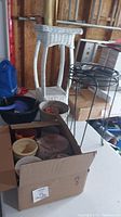 Overview photo showing white wicker plant stand, black metal plant stand, assorted plant pots, and cardboard box containing pots and watering can
