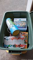 Green storage tote containing multiple boxed jigsaw puzzles and one cylindrical puzzle box.