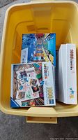 Top-down photo of four visible puzzle boxes inside a yellow plastic tote; boxes show colorful 1,000-piece puzzles including a canal street scene and a photo collage theme