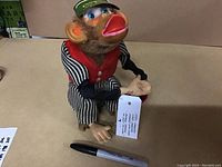 Front angle showing monkey toy sitting on beige surface holding a tag, with a black marker for size reference. The toy wears a red vest and black and white striped pants and shirt with detailed facial features and fur accents.