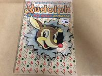 The front cover of the 1960 Rudolph the Red Nosed Reindeer comic book, showing colorful illustration of Rudolph breaking through a decorated background with repeating holiday motifs. Comic is inside a clear protective plastic sleeve with light reflections.