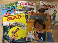 Photo showing five vintage magazines laid out with colorful covers: two MAD magazines and three Cracked magazines (including one Collector's Edition). Covers depict caricatures, comic themes, and pop culture references.