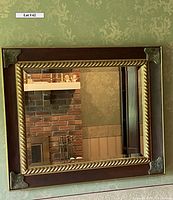 Frontal photo showing rectangular mirror with dark wood frame, gold rope accent and ornate metal corner pieces mounted on wall covered in patterned wallpaper. Mirror reflects part of brick fireplace and shelf.