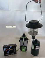 Full lot shot with Coleman camping lantern held by handle, Coleman water bottle in black, gray, and green sleeve, new unopened Coleman propane tank, and boxed solar charger.