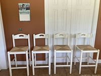Four white wooden counter height stools with woven rush seats in a row showing overall condition and size.