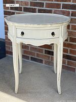 Creamy white vintage round accent table with one drawer, showing front angle with minor scratches and paint wear.