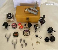 Overview photo of the lot including five fishing reels, yellow tackle box, metal fish scaler, fishing lines, bobbers, sinkers, hooks, and lure components all arranged on a flat surface.