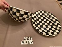 Photo showing the 11-inch round bowl held and the 16-inch round platter on a surface, both with a black and white checker pattern and gold rims.