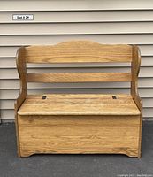 Front view of a solid oak storage bench with a hinged lift-top seat and slatted backrest, placed against an exterior wall with vinyl siding.