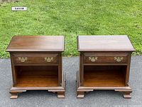 Pair of solid cherry nightstands photographed outdoors on pavement with grass background, showing front view, each with one drawer and open shelf below.
