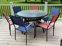 Photo showing complete outdoor dining set with round rattan table, glass top, and six metal mesh chairs in red and blue, taken on a deck