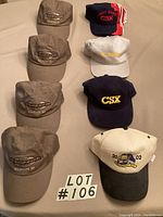 Eight CSX hats arranged in two columns on beige background including four brown Albany Division hats, tan 2002 Rochester NY hat, black hat, white hat with yellow CSX logo, and navy blue Albany Division hat with red/white stripes