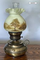 Vintage electric Fenton lamp with a hand-painted glass shade showing landscape scenes in brown on creamy-white glass. The lamp has a clear glass chimney and tarnished metal base with a curved handle.
