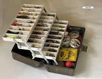 Open metal tackle box showing multiple fold-out trays filled with assorted fishing lures and accessories