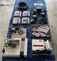 Full lot of vintage gaming items laid out on blue cloth including Nintendo NES console, NES Advantage joystick controller, four NES game cartridges, Nintendo light zapper, various AV and power cables, and boxed Retron FC Loader console with two controllers.