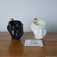 Front view of two ceramic teacups shaped as baby doll heads in black and white with gold handles sitting on a wooden surface.