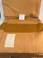 Folded wool blanket displaying the large label with gold text "The Seal of Quality, 100% Wool Laine" and tan and brown colored fabric.