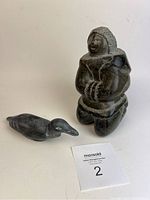 Photo showing the Inuit soapstone sculptures of a woman in a parka and a bird on a white background with a lot number card.