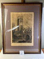 Framed engraving showing tall chapel tower by river with stone bridge and rowboat below. Mounted with matting and glass in a wooden frame with gold inset.