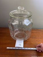 Full view of the square clear glass jar with embossed Planters Peanuts logo and a measuring tape indicating about 12 inches tall.