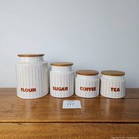 Full set of 4 ceramic canisters labeled FLOUR, SUGAR, COFFEE, TEA with wooden lids on a wood table