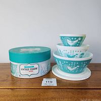 Front view of three nested turquoise decorated opal glass mixing bowls with white plastic lids next to vintage round hat box with Vintage Charm Pyrex branding.