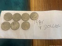 Seven Canadian one-dollar coins from 1985 laid out on a white napkin with handwritten note reading '1985 7 Dollar'. Obverse side with Queen Elizabeth II profile visible.