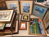 Photo showing assortment of framed art including harbor scenes, rural houses, and floral still life, stacked and arranged on table