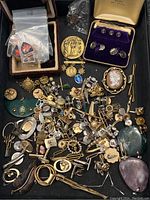 Overhead view of black tray holding mixed jewelry components: cufflinks, tie bars, pins, medal, cameo and stone pendants
