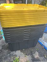 Photo showing stacked plastic storage totes with yellow lids on top and black bases beneath, outdoors on pavement.