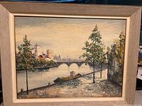 Front view of framed oil on canvas painting showing riverside scene with bridge, trees, and boats.