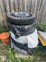 Stack of four tires outdoors by wooden fence with two wrapped in black and white plastic bags and one on top showing black steel spare rim with speed limit labels.