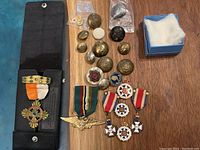 Overall view of medals, buttons, pins including one medal with orange and white ribbon and star-shaped cross in black case, assorted buttons with various insignia.