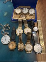 Overview of vintage wristwatches and pocket watch laid out on surface, showing variety of bands and dial conditions.