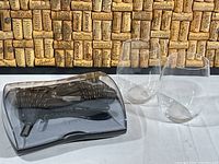 Shows Metrokane Rabbit corkscrew in smoky gray plastic storage case along with two clear stemless Riedel wine glasses in front of a wine cork backdrop