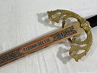 Close-up photo of sword blade showing red and black engraving with 'TIZONA DEL CID' inscription and ornate gold-colored guard.