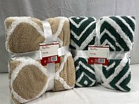Two Manta Sherpa throw blankets in original packaging, one tan/white circular pattern and one dark green/white chevron pattern, tied with a white ribbon.