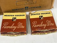 Two Webster-Chicago labeled recording wire boxes in brown and yellow, one slightly damaged with a metal wire reel inside.