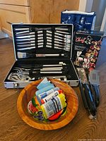 Image showing full BBQ set arranged with wood fruit bowl containing ice packs and cooler bag behind. The aluminum case is open displaying set of stainless steel utensils and Master Chef Grill Caddy box.